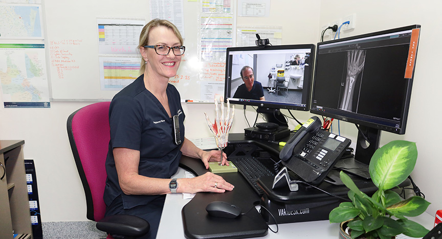 A healthcare professional sits at a desk with two computer monitors displaying a video call and an X-ray image. A model of a hand is on the desk.