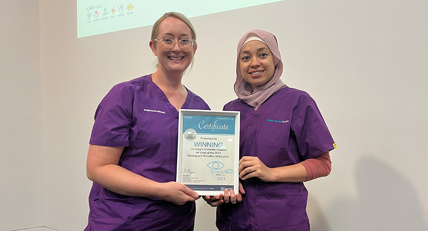 Two healthcare workers in purple uniforms hold a certificate for winning the Nursing and Midwifery Graduate award.