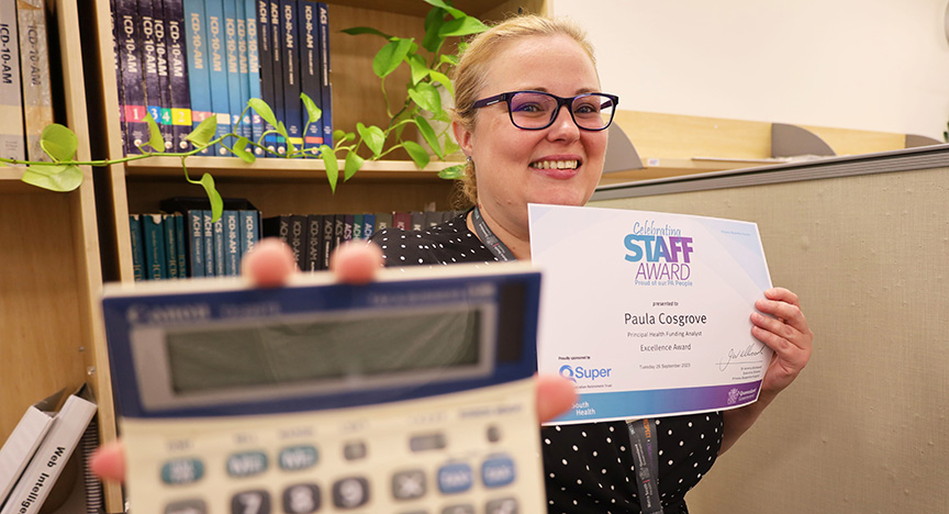 A person holds a certificate for an Excellence Award in one hand and a calculator in the other, standing in front of a bookshelf.