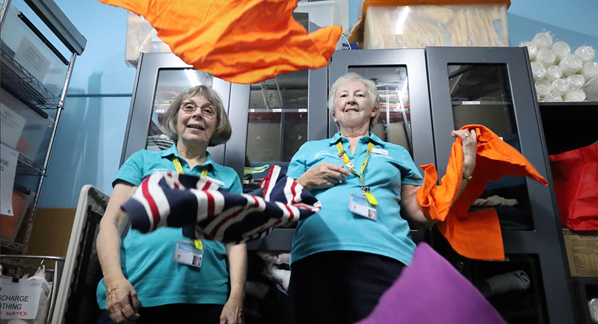 Two volunteers in blue shirts sort and fold colorful clothing in a storage room.