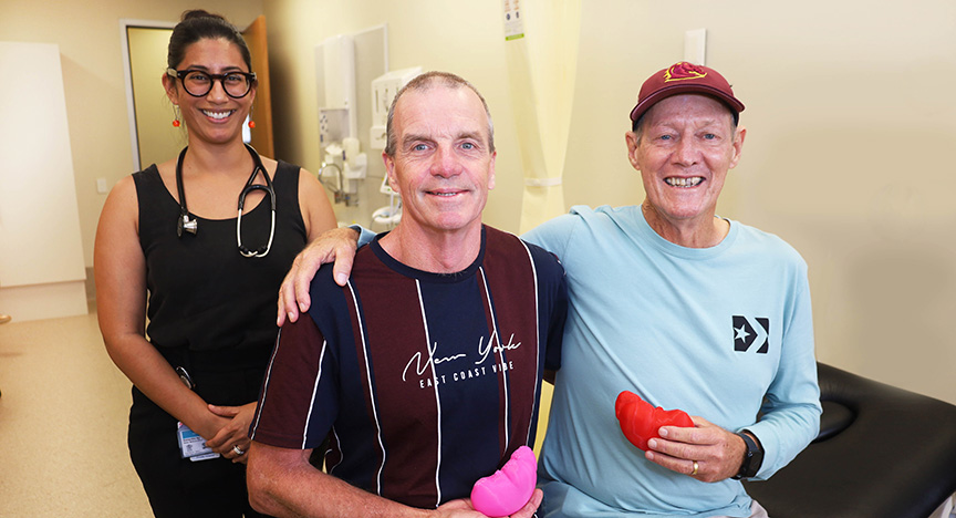 Two people sit in a medical setting holding red and pink kidney-shaped objects, with a healthcare worker standing behind them.