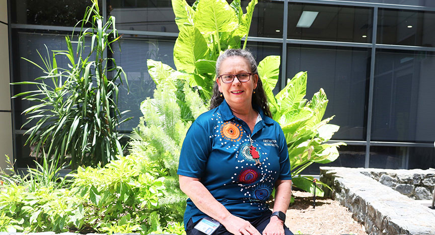 A person in a First Nations Health shirt sits in a garden with large green plants, in front of a building with glass windows.