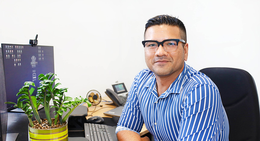 A person in a blue striped shirt sits at a desk with a computer, phone, and a potted plant.