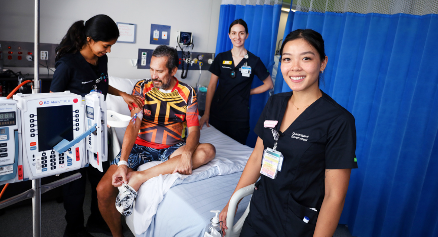 young nurses in scrubs at the bedside tending to a male patient wearing an indigenous shirt. An experienced nurse overseas care