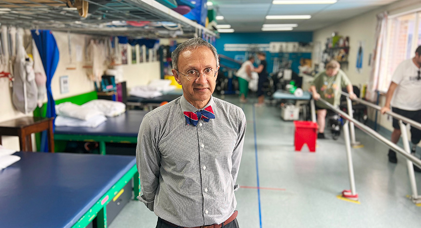 A person in a striped shirt and bow tie stands in a rehabilitation center with equipment and people exercising in the background.