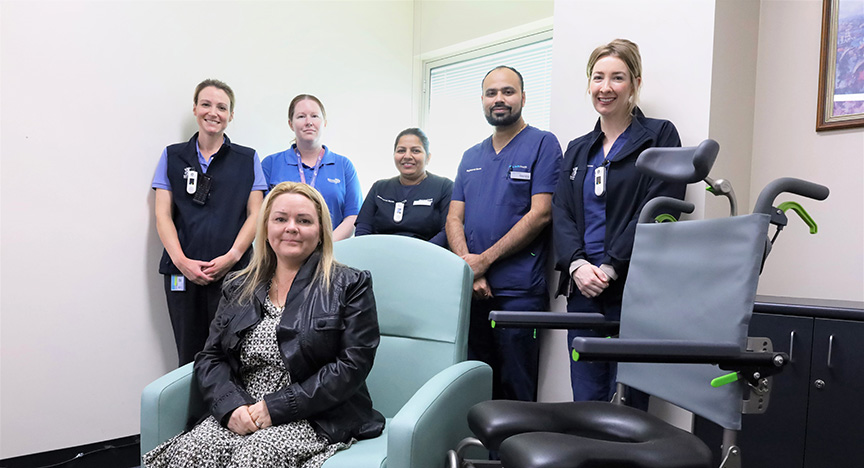 A person sits in a chair, surrounded by five healthcare workers standing. A medical chair is beside them.