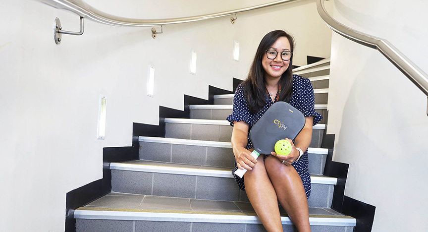 A person sits on stairs holding a pickleball paddle and ball. The setting appears to be indoors with a curved handrail.