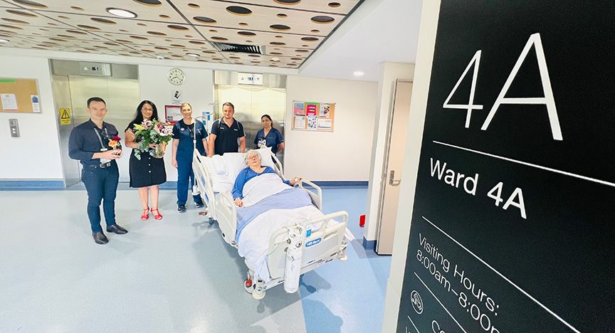 A patient in a hospital bed is surrounded by staff and visitors in Ward 4A. A sign shows visiting hours from 8:00 am to 8:00 pm.