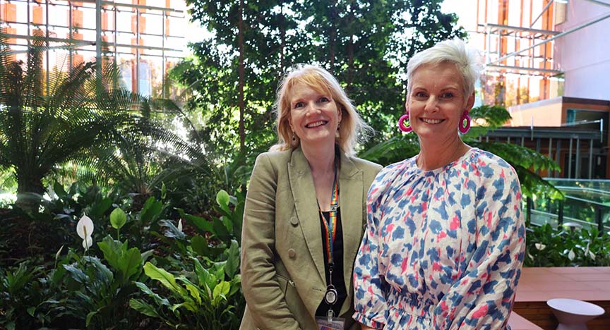 Two people standing in a lush indoor garden with large windows in the background. One wears a green blazer, the other a colorful dress.