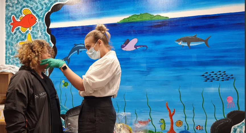 A healthcare worker examines a person's mouth in front of a colorful ocean-themed mural.