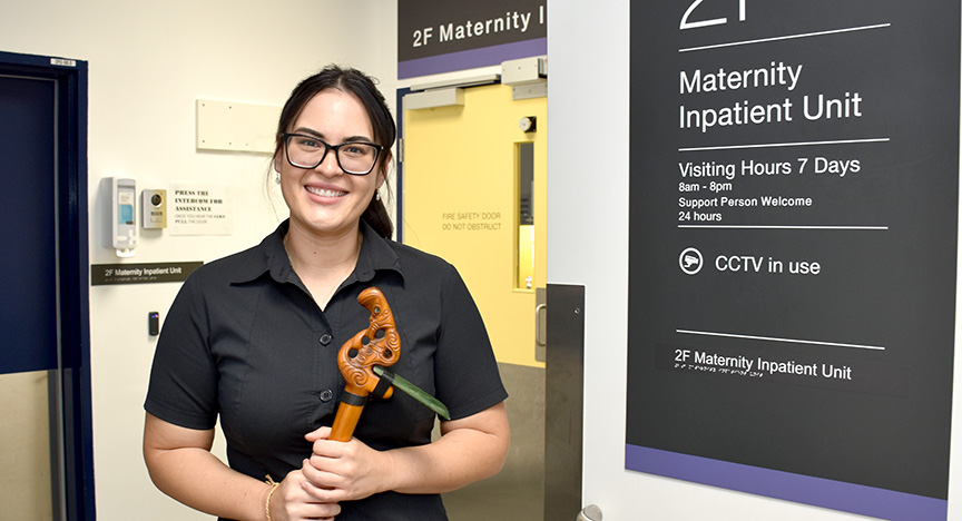 A person holding a carved wooden object stands in front of the 2F Maternity Inpatient Unit sign, which lists visiting hours and CCTV usage.