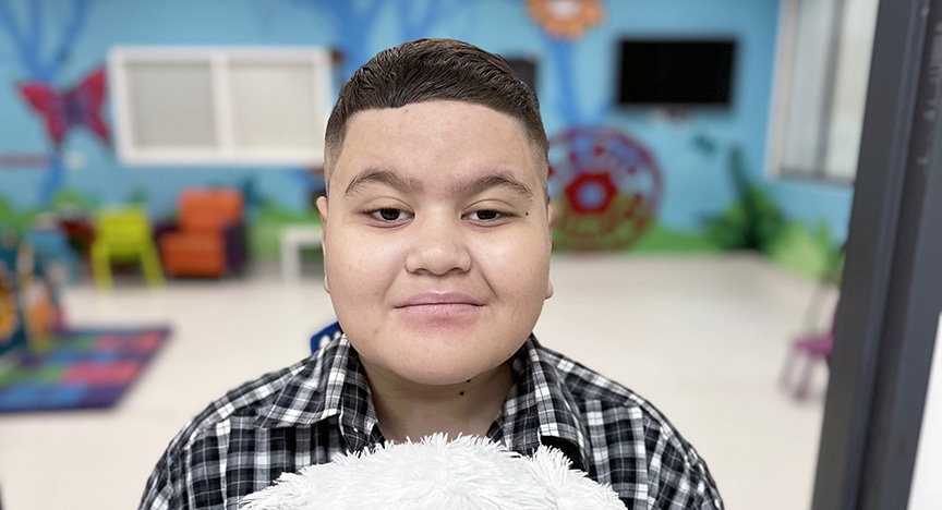 A person in a checkered shirt holds a white stuffed animal in a colorful room with chairs, a TV, and wall decorations.