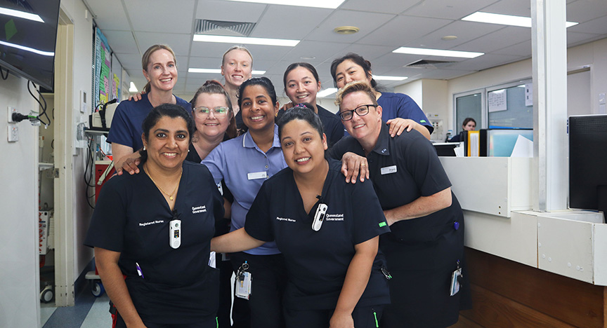 A group of healthcare workers in uniforms stands together in a hospital ward, smiling at the camera.