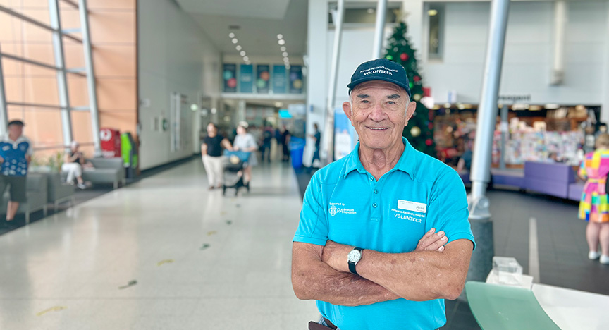 A volunteer in a blue shirt and cap stands with arms crossed in a hospital lobby. People and a Christmas tree are visible in the background.