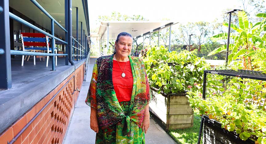 A person stands on a pathway between a building and a garden with lush green plants. The person is wearing a colorful outfit.