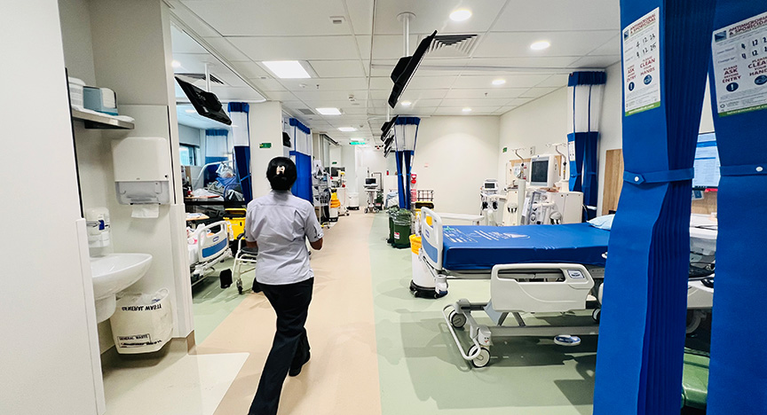 A healthcare worker walks through a hospital ward with multiple beds and medical equipment.