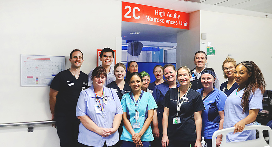 A group of healthcare workers stands in front of the entrance to the 2C High Acuity Neurosciences Unit in a hospital.