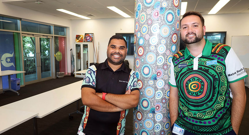 Two people in colorful shirts with First Nations designs stand beside a decorated pillar in a modern room with large windows.