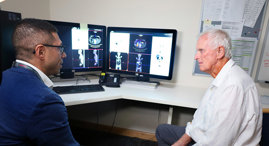 A doctor and a patient review medical scans on computer monitors in a clinical setting.