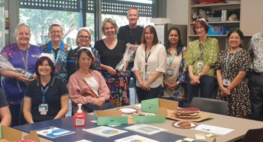 A group of people standing in a room with tables displaying cakes, documents, and hand sanitizer, celebrating International Nurses Day.