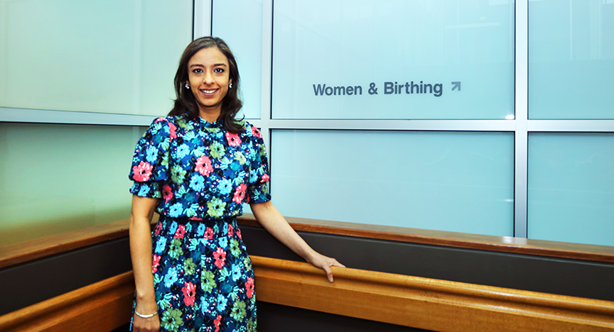 A person in a floral dress stands near a sign that reads Women & Birthing in a hospital setting.