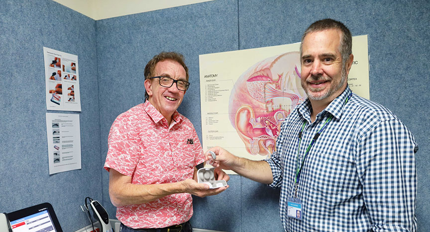 Two people in a medical office holding a hearing aid model, with an ear anatomy poster in the background.