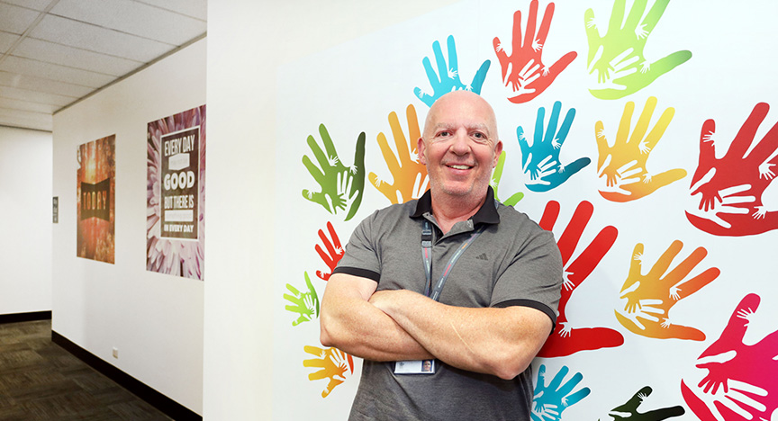 A person stands with arms crossed in front of a wall decorated with colorful handprints. Posters with motivational messages are on the hallway wall.