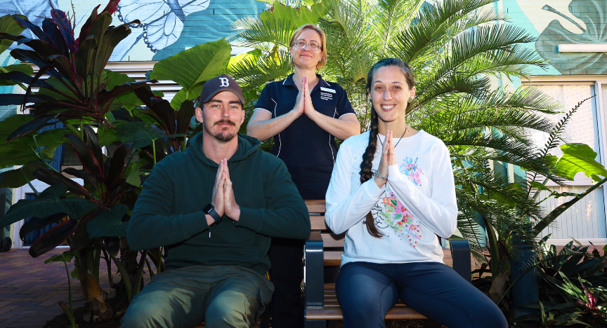 Three people practicing yoga in a garden, sitting on a bench with hands in prayer position, surrounded by lush green plants.