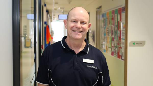 A physiotherapist stands in a hospital hallway, wearing a name tag and a black polo shirt with PHYSIOTHERAPY written on it.