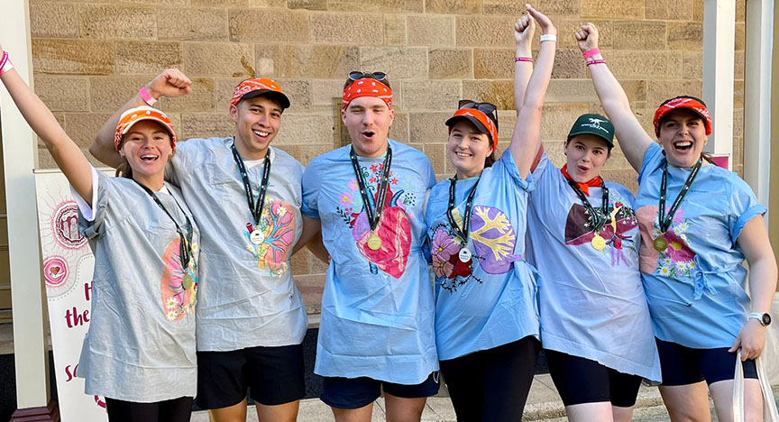 A group of people wearing matching shirts and medals, celebrating with raised arms in front of a brick wall.