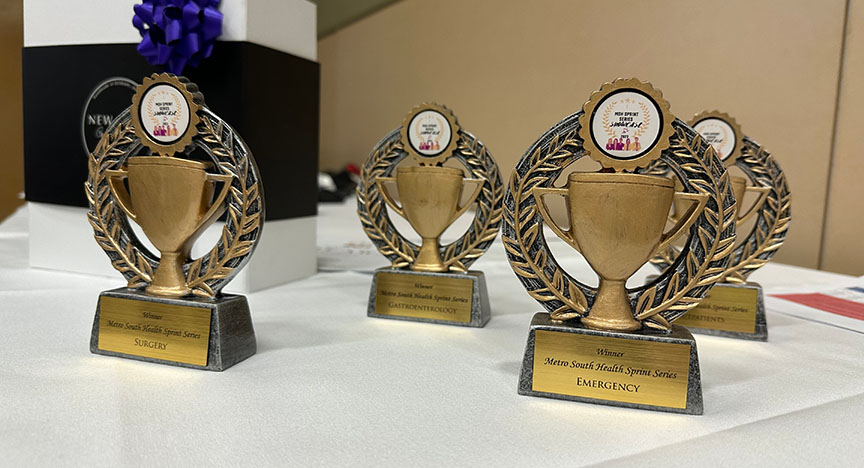 Four gold trophies with laurel wreaths on a table, each labeled with different medical specialties: Surgery, Gastroenterology, and Emergency.