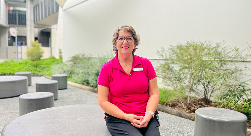 A person in a pink shirt with a name tag sits on a round concrete bench in an outdoor area with greenery and modern architecture.