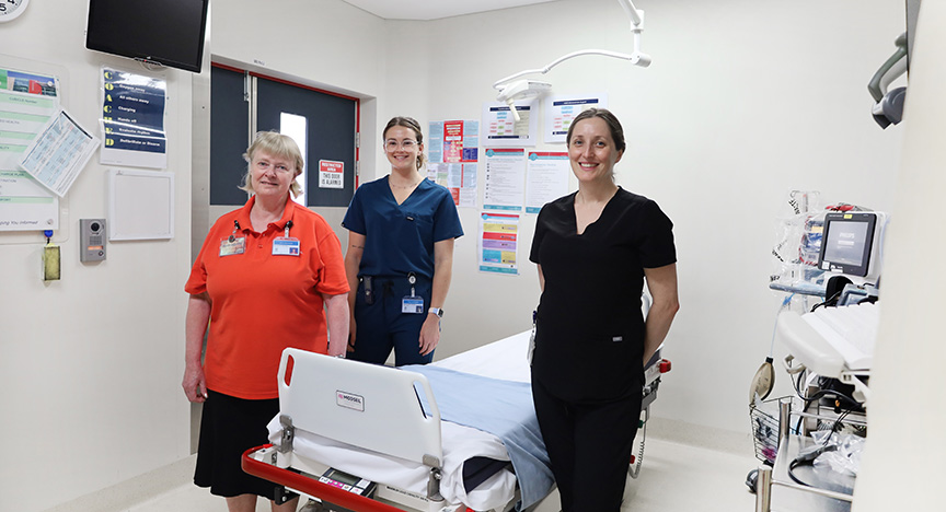 Three healthcare workers stand in a hospital room with medical equipment and a bed. They are wearing uniforms and name badges.