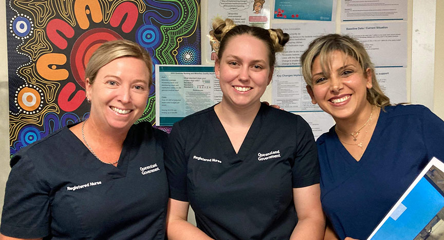 Three registered nurses in Queensland Government uniforms stand in front of a First Nations artwork and informational posters.