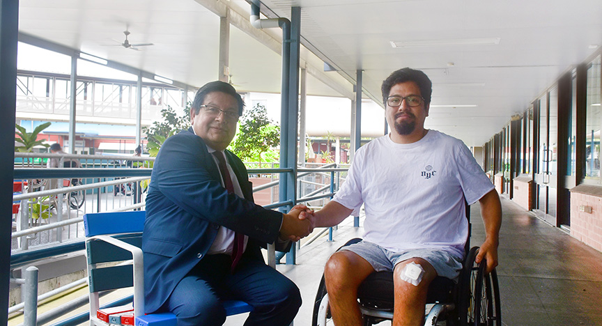 A person in a suit shakes hands with a person in a wheelchair on a covered walkway. The person in the wheelchair has a bandage on their knee.