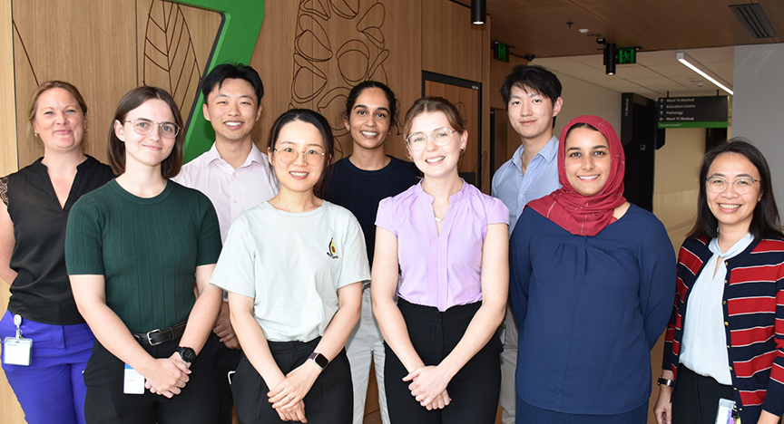 A group of nine students standing indoors, some wearing ID badges. The background features wooden walls with leaf designs.