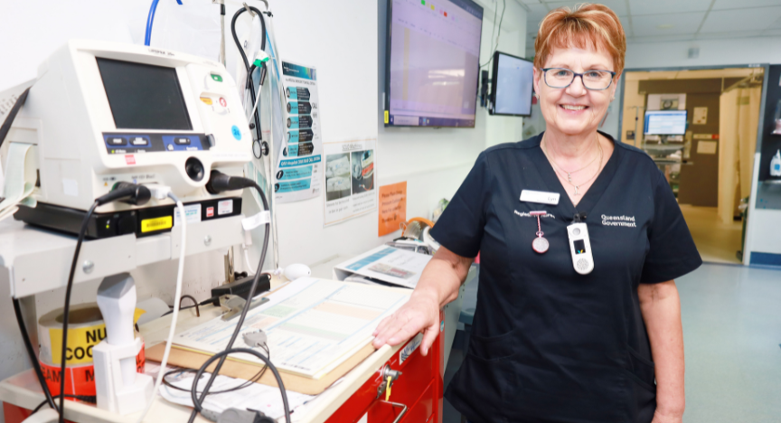 A nurse stands beside medical equipment in a hospital room, wearing a Queensland Government uniform.