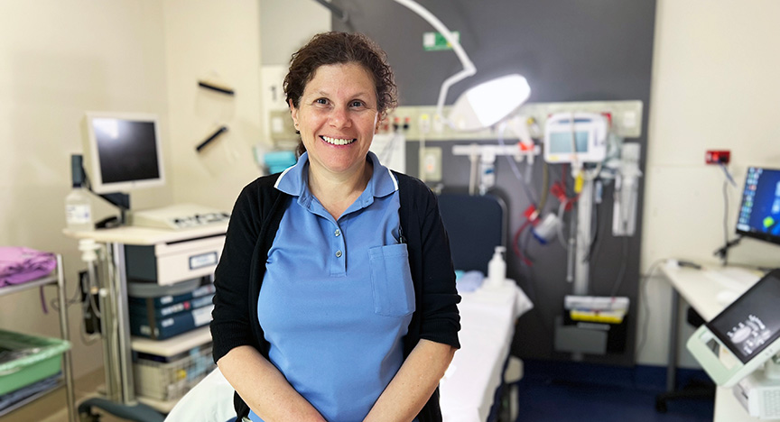 A nurse in a blue uniform stands in a medical room with various equipment and monitors.