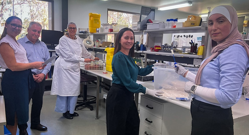 A group of people in a laboratory, some in lab coats and gloves, working with scientific equipment and documents.