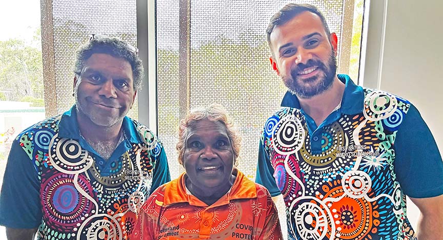 Three people wearing colorful shirts with First Nations designs stand together indoors. The middle person wears an orange shirt with COVID PROTECT text.