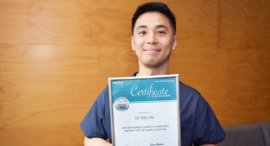 A person in scrubs holds a Certificate of Achievement awarded to Dr. Ken Ho for qualities of collaboration, teamwork, and high-quality clinical care.