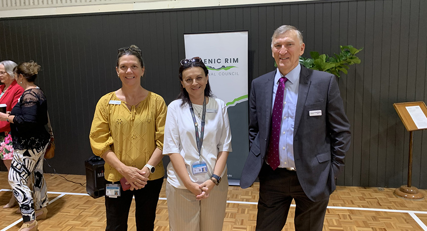 Three people stand indoors in front of a Scenic Rim Regional Council banner. Other people are in the background.