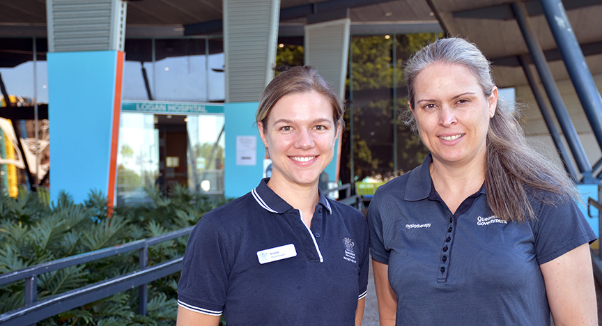 Two healthcare workers in uniform stand outside a hospital entrance. One wears a name badge, and the other has physiotherapy on their shirt.