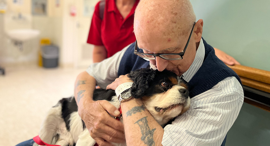 An elderly person with tattoos hugs a small black and white dog in a hospital setting. Another person in a red shirt stands nearby.