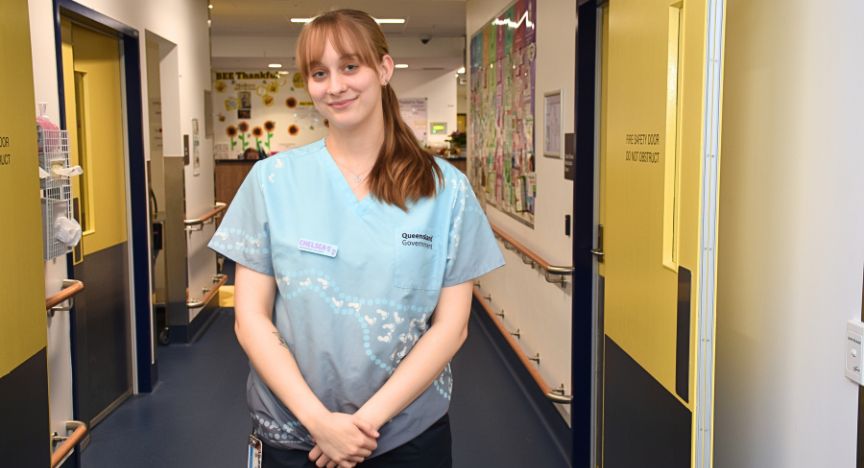 A healthcare worker in a blue uniform stands in a hospital corridor. The uniform has a Queensland Government logo and a name badge.