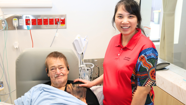 A patient in a hospital bed is visited by a person in a red shirt with First Nations artwork. Medical equipment is visible in the background.