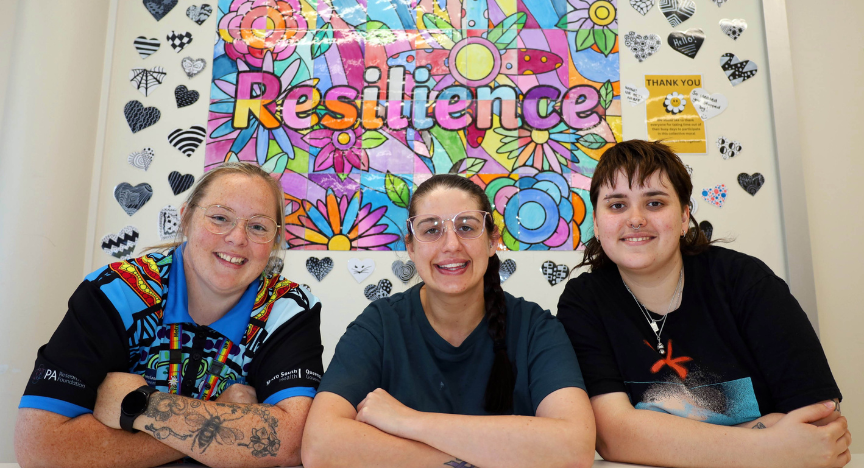 Three peer workers sitting with elbows on table in front of colourful mural which spells with word 'resilience'