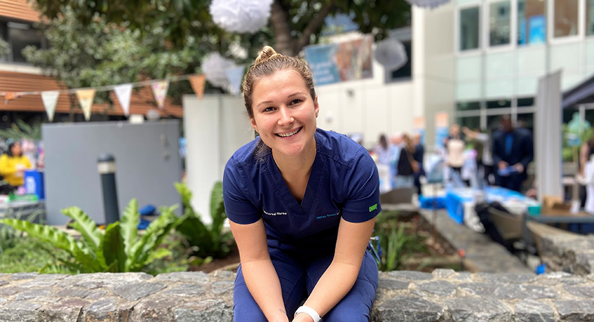 A person in blue scrubs sits on a stone ledge outdoors at a community event with people and booths in the background.