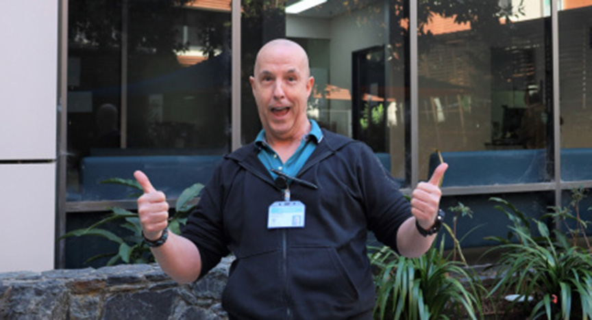 A person stands outside a building, giving two thumbs up. They are wearing a lanyard with an ID badge. There are plants and windows in the background.