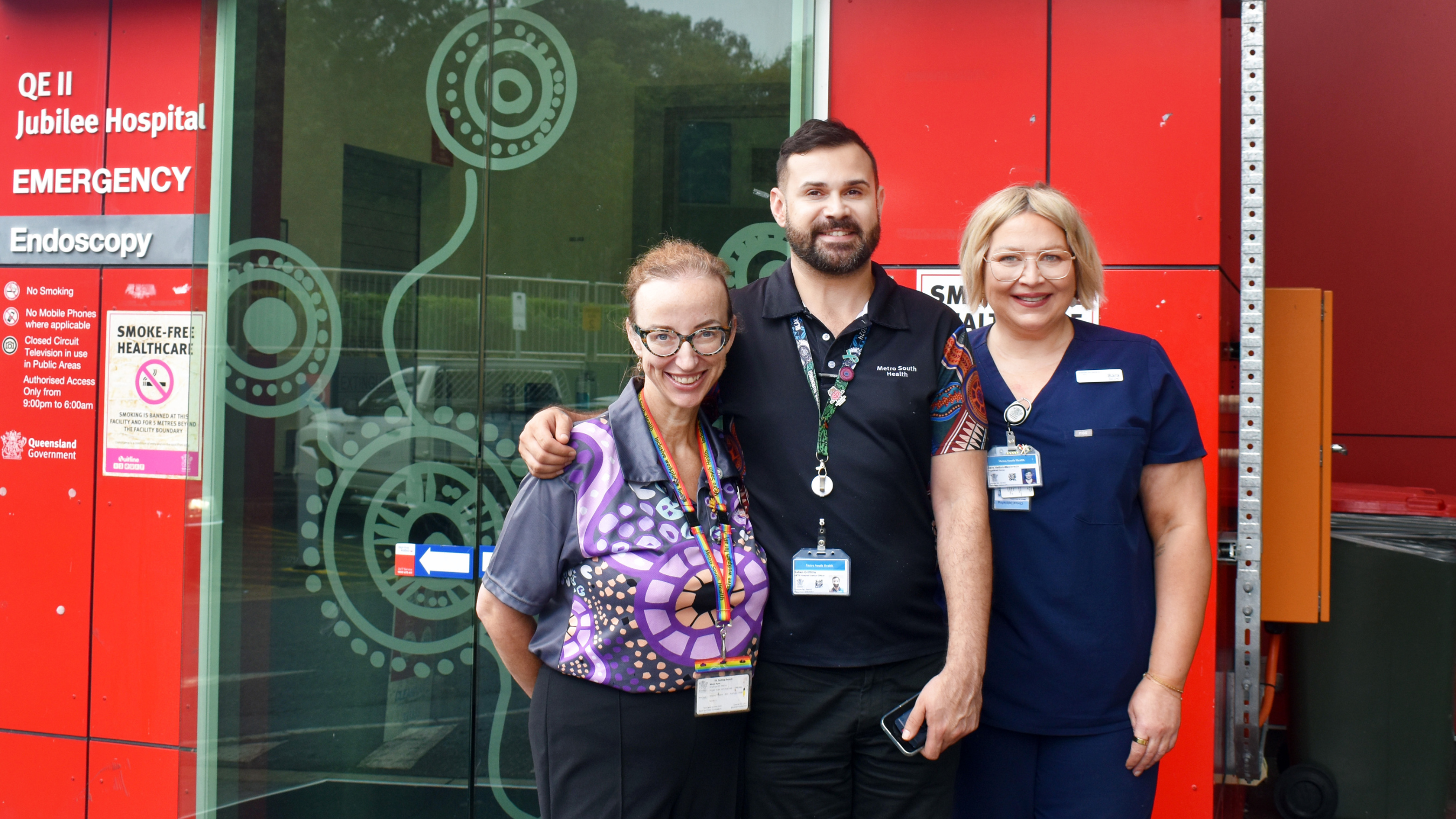 3 staff members standing outside QEII Emergency entrance with glass doors displaying songlines art